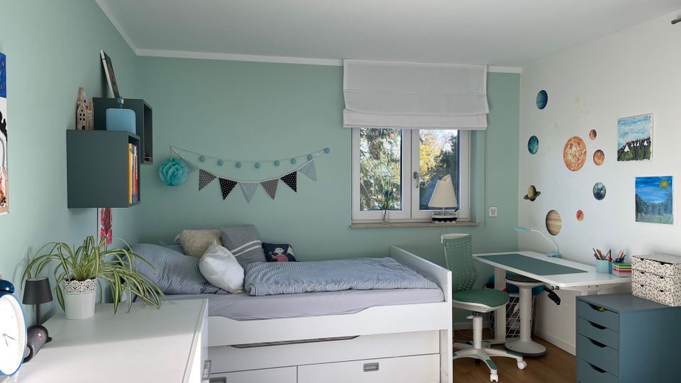 Helles Kinderzimmer mit mintgrünen Wänden, einem weißen Einzelbett mit Schubladen, einem Schreibtisch mit grünem Stuhl und einer Kommode. Die Wand über dem Bett ist mit einer Wimpelkette dekoriert, während die gegenüberliegende Wand ein Planetensystem und Bilder zeigt; ein Fenster mit Blick auf Bäume lässt Tageslicht herein.