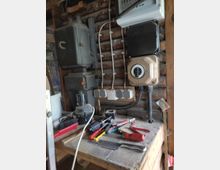 Eine Werkbank in einer Holzwerkstatt mit verschiedenen Werkzeugen wie Schraubendrehern, Zangen und einer Säge auf der Arbeitsfläche. Im Hintergrund befinden sich Holzvertäfelungen, elektrische Anschlüsse und Sicherungskästen.