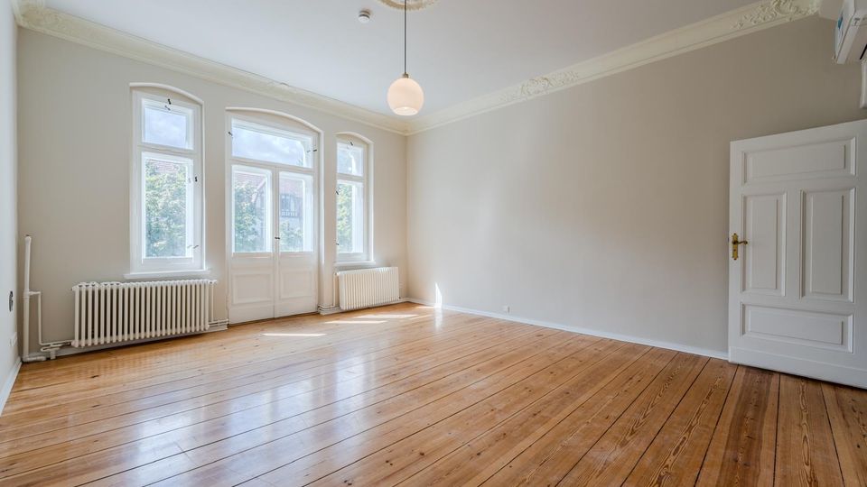 Heller, leerer Altbau-Wohnraum mit hohen Decken, Stuckverzierungen und einem runden Deckenleuchter. Drei hohe Fenster und eine verglaste Flügeltür lassen viel Tageslicht auf den Holzdielenboden; an den Wänden stehen Heizkörper und eine weiße Zimmertür ist rechts sichtbar.