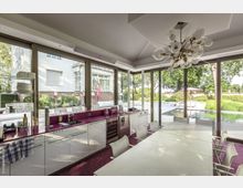 Moderner Innenraum einer Küche mit großen bodentiefen Fenstern, die den Blick auf einen gepflegten Garten mit Pool und Terrasse freigeben. Die Küche verfügt über eine Arbeitsplatte in kräftigem Pink, helle Möbel und eine auffällige Deckenleuchte.