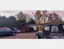 Ein Werkstatthof mit geparkten Autos, darunter ein Cabrio auf einem Container und mehrere Fahrzeuge, inklusive eines Jeeps mit Verdeck. Im Hintergrund sind Bäume und ein flacher gelber Gebäudeteil zu sehen, vor einem Abendhimmel mit rötlichem Licht.
