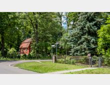 Eine ländliche Szene mit einem kleinen roten Haus mit Satteldach, umgeben von hohen Bäumen und dichtem Grün. Im Vordergrund befindet sich eine Straße mit einer Kurve, ein Metallzaun und ein Straßenschild.