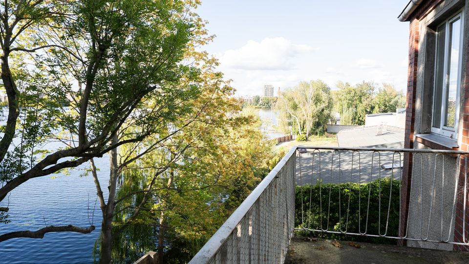 Blick von einem kleinen Balkon an einem Backsteingebäude mit Metallgeländer auf einen See, der von dichtem Baumbestand gesäumt ist. Im Hintergrund sind ein Uferbereich mit weiteren Gebäuden und ein entferntes Hochhaus unter hellem Himmel zu sehen.
