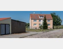 Mehrfamilienhaus mit vier Wohneinheiten und rotem Ziegeldach in ländlicher Umgebung, daneben ein einfacher Garagenkomplex mit flachem Dach und grauen Türen; im Vordergrund eine gepflasterte Straße und ein Gehweg.