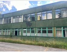 Zweistöckiges, längliches Gebäude mit dunkelgrüner Fassade und Reihen aus großen Fenstern mit grünen Rahmen, teils mit Vorhängen. Vor dem Gebäude verläuft ein Betonweg mit hohem, verwildertem Gras am Rand; darüber ist ein teilweise bewölkter Himmel zu sehen.