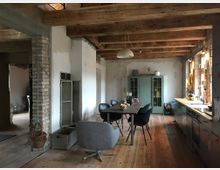 Rustikaler Wohnraum mit offener Küche, Holzbalkendecke und Holzboden. Im Raum stehen ein Esstisch mit Stühlen, eine Metallküchenzeile und ein blaues Schrankmöbel; sichtbare Wände sind teils unverputzt oder aus Ziegeln.