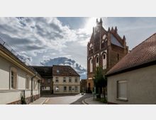 Kleinstadt-Straßenszene mit einer historischen Backsteinkirche im gotischen Stil im Hintergrund. Die Umgebung zeigt Wohnhäuser mit roten Ziegeldächern und gepflasterte Gehwege unter bewölktem Himmel.