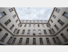 Innenhof eines mehrstöckigen, historischen Gebäudes mit heller Fassade und zahlreichen symmetrisch angeordneten Fenstern. Der Himmel ist bewölkt, und an einer Wand ist eine Klimaanlage befestigt.