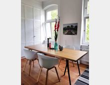 Esszimmer mit hellen Holzböden und einem einfachen rechteckigen Holztisch, um den vier weiße Stühle stehen. Große weiße Fenster lassen Tageslicht herein, und an der Wand hängt ein Landschaftsbild. Eine Vase mit roten Blumen und dekorative Gegenstände stehen auf dem Tisch.