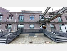 Backsteinfassade eines historischen Industriegebäudes mit großen grünen Fenstern und Türen. Vor dem Gebäude befinden sich Treppen und ein Metallsteg, darüber verlaufen metallene Konstruktionselemente einer ehemaligen Förderanlage.