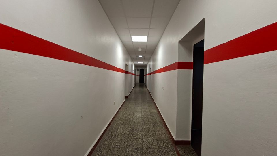 Langer, schmaler Innenflur mit weißen Wänden und einem durchgehenden roten Querstreifen auf beiden Seiten. Grauer Steinboden, abgehängte Decke mit rechteckigen Deckenleuchten und mehrere Türöffnungen entlang des Gangs.