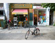 Straßenszene in einer urbanen Umgebung mit zwei kleinen Geschäften: links ein Laden mit der Aufschrift „Koof im Kiez!“ und roter Markise, rechts ein Geschäft mit dem Namen „East Berlin“ und ausgestellten Kleidungsstücken. Vor den Läden stehen Fahrräder, und Passanten bewegen sich in der Szene.