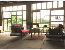 Industriehalle mit großen Fensterfronten, durch die eine grüne Landschaft sichtbar ist. Im Vordergrund stehen zwei Metallbetten mit roten Polstern, ein Tisch mit weißer Tischdecke und ein Kleiderständer; der Boden ist roh und uneben.