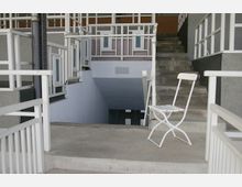 Ein moderner Gebäudeeingang mit weiß gestrichenen Geländern, Betonstufen und einer Treppe, die in einen unteren Bereich führt. Ein einfacher weißer Stuhl steht auf einer Betonplattform im Vordergrund.