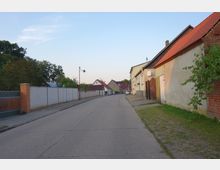 Ländliche Straße mit einfachen Häusern, einige mit roten Ziegeldächern, und einer weißen Mauer auf der linken Seite. Die Umgebung enthält Bäume und grüne Flächen, während eine Laterne und ein geparktes Auto im Hintergrund sichtbar sind.