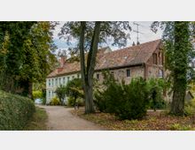 Historisches Gebäude mit Fachwerk- und Steinwänden sowie einem roten Ziegeldach, umgeben von Bäumen und Sträuchern in einem bewaldeten Parkbereich mit einem verschlungenen Kiesweg im Vordergrund.