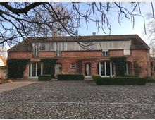 Backsteingebäude mit begrünten, bogenförmigen Eingängen und großen Fenstern, umgeben von einem gepflasterten Hof. Im Vordergrund kahle Äste eines Baumes und geometrisch geschnittene Hecken.