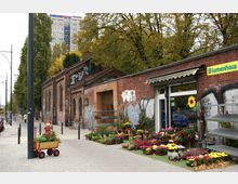 Eine Straßenszene mit einem Backsteingebäude, das mit Graffiti versehen ist, und einem Blumengeschäft mit bunten Blumenarrangements vor dem Eingang. Im Hintergrund sind Bäume und ein Wohnhochhaus zu sehen.