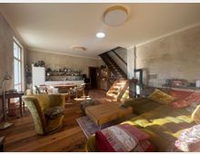 Wohnbereich in einem offenen Raum mit rustikalem Design, Holzboden und beigen Wänden. Im Vordergrund steht ein großes Sofa mit bunten Kissen, daneben ein runder Esstisch mit Stühlen und eine offene Holztreppe, die zu einem oberen Stockwerk führt.