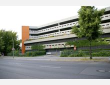 Mehrstöckiges Parkhaus mit weißen und orangefarbenen Fassadenelementen, teilweise von Efeu bewachsen. Im Vordergrund eine Straße mit Gehweg, Schildern und einem Baum.