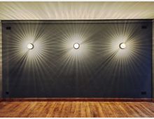 Dunkler Raum mit einer grauen Wand, an der drei symmetrisch platzierte Wandleuchten befestigt sind. Die Lampen strahlen jeweils kreisförmige Lichtmuster, und der Boden besteht aus hellem Holz.