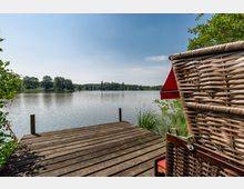 Holzsteg an einem ruhigen See mit umliegender Ufervegetation und Wald am Horizont; am rechten Bildrand ist ein Strandkorb mit rotem Sonnendach sichtbar.