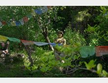 Eine Gartenlandschaft mit Bäumen und dichtem Grün, in dessen Zentrum eine steinerne Statue einer Figur steht. Gebetsfahnen in verschiedenen Farben hängen quer über den Bereich gespannt.