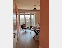 Ein moderner Wohnraum mit hellrosa gestrichenen Wänden und hellbraunem Holzboden. Im Vordergrund steht ein Holztisch mit Stühlen, daneben eine Tüte auf dem Boden, im Hintergrund eine große Fensterfront mit Zugang zu einem Balkon und Blick auf Gebäude in der Ferne.