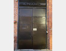 Große, dunkle Metalltür mit der Aufschrift „REINGOLD“ oben über der Tür. Die Tür ist in eine rote Ziegelwand eingebaut, und ein kleiner Hinweis mit der Aufschrift „Please Ring the Bell“ ist in der Mitte der Tür angebracht.