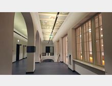 Heller Innenflur eines historischen Gebäudes mit hohen Rundbögen, großen Sprossenfenstern und einem zentralen Treppenabgang mit Geländer. An der Decke verläuft eine lange Lichtschiene unter bemalten Kassettfeldern mit dekorativen Motiven.