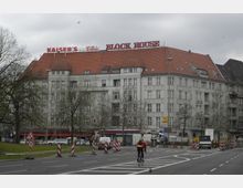 Mehrstöckiges Eckgebäude mit heller Fassade und rotem Ziegeldach, auf dem rote Leuchtschrift mit den Worten „Kaiser’s“ und „Block House“ steht. Im Vordergrund verläuft eine Straße mit Baustellenabsperrungen, Verkehrszeichen und einer Radfahrerin auf der Fahrbahn.