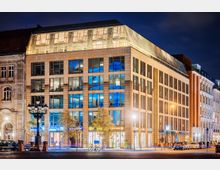 Moderne Büro- und Geschäftsgebäude bei Nacht mit großer Glasfassade, beleuchteten Fenstern und Geschäften im Erdgeschoss, links angrenzend an ein historisches Gebäude mit verzierten Fassaden. Im Vordergrund eine Straßenlaterne und parkende Autos.