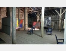 Innenraum einer rustikalen Lagerhalle mit Holzbalken und Holzboden, genutzt als Kunstausstellungsraum. Mehrere abstrakte Gemälde sind auf Staffeleien und Wänden ausgestellt, umgeben von einigen Kunststoffstühlen.