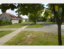 Straßenszene in einem Dorf mit freistehenden Häusern, teils mit roten Ziegeldächern, entlang einer kleinen Straße. Im Vordergrund Gehwege und grüne Rasenflächen, im Hintergrund Bäume und weitere Häuser.