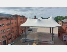 Ehemaliges Industriegebäude aus rotem Backstein mit kleinen, symmetrischen Fenstern umgibt einen gepflasterten Platz mit einer großen, weißen, zeltartigen Überdachung auf Metallstützen. Im Hintergrund sind Bäume und ein blauer Himmel sichtbar.