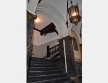 Treppenhaus in historischem Gebäude mit schwarzen Marmorstufen, verzierten weißen Bögen, goldenen Dekorationen und einer großen, hängenden Laterne. Im Hintergrund ist eine Holzbalustrade zu sehen.
