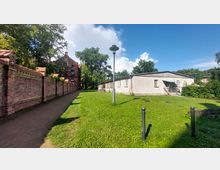 Wegezugang mit einer roten Backsteinmauer auf der linken Seite, einem historischen Backsteingebäude im Hintergrund und einer einfachen weißen Betonstruktur auf der rechten Seite. Im Vordergrund befindet sich eine Grünfläche mit einer Laterne in der Mitte.