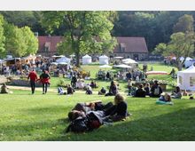Grüne Parklandschaft mit Menschen, die auf der Wiese sitzen oder liegen, im Hintergrund ein Marktplatz mit weißen Zelten und Ständen sowie ein langes Gebäude mit rotem Ziegeldach. Umgeben von Bäumen und Wald wirkt die Szene naturnah und belebt.