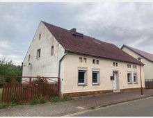 Ein einstöckiges Wohnhaus mit beigefarbener Fassade, roten Dachziegeln und mehreren rechteckigen Fenstern. Vor dem Haus befindet sich ein kleiner Zaun, und die Straße ist mit Gehwegplatten ausgelegt.