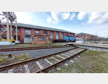 Ansicht eines Eisenbahndepots mit einem roten Backsteingebäude im Hintergrund, einer blauen Lokomotive auf einer Drehscheibe und mehreren Eisenbahnschienen im Vordergrund. Rechts sind weitere Backsteingebäude und Vegetation sichtbar, der Himmel ist leicht bewölkt.
