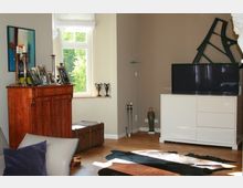 Wohnzimmer mit einer Mischung aus modernen und klassischen Möbeln. Im Vordergrund ein Holztisch und ein beigefarbener Sessel mit lila Kissen; im Hintergrund eine Holzkommode mit Bilderrahmen und Kerzenständern, ein weißer Fernsehtisch mit Fernseher und eine große Fensterfront, durch die Tageslicht einfällt.