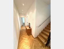 Schmaler, heller Flur in einem Wohnhaus mit weißen Wänden, eingelassenen Deckenleuchten und Fischgrät-Holzboden. Rechts führt eine Holztreppe mit Metallhandlauf nach oben, links stehen Türen zu angrenzenden Räumen und ein niedriger Holzschrank an der Wand.