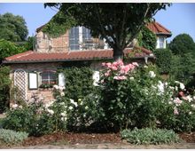 Backsteinhaus mit rotem Ziegeldach, umgeben von blühenden Rosensträuchern und Kletterpflanzen, im Hintergrund dichter Baumbestand. Ein Baum steht im Vorgarten, und eine kleine Terrasse ist im oberen Stockwerk sichtbar.