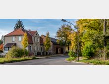 Ecke einer Dorfstraße mit einem zweistöckigen Wohnhaus mit rotem Ziegeldach links und einer großen Scheune aus Backstein im Hintergrund. Der Bereich ist von Bäumen und Herbstlaub umgeben, mit einem Gehweg und einer Straßenlaterne rechts.