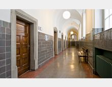 Ein langer, gewölbter Flur in einem historischen Gebäude mit Türen aus Holz und Wänden, die teilweise mit glänzenden, dunklen Fliesen verkleidet sind. Der Boden besteht aus poliertem Stein, und kugelförmige Deckenlampen beleuchten den Raum; an einer Seite befinden sich Sitzbänke und ein Heizkörper unter einem Fenster.