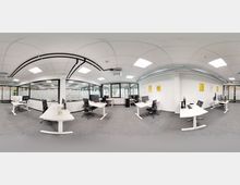 Moderner Büroraum mit mehreren weißen Schreibtischen und schwarzen Bürostühlen, ausgestattet mit Monitoren und Büroartikeln. Große Fenster mit Sichtschutzfolien und Glaswänden trennen den Raum, während sichtbare Lüftungsrohre und Deckenbeleuchtung die minimalistischen Elemente unterstreichen.