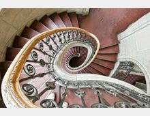Spiralförmige Treppe mit roten Stufen und kunstvollem Metallgeländer, die in einem historischen Gebäude hinunterführt. Die geschwungenen Formen des Geländers und die gleichmäßige Wiederholung der Stufen betonen die spiralförmige Architektur.