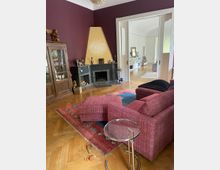 Wohnzimmer mit Holzboden und bordeauxroten Wänden. Im Vordergrund steht ein rotes Sofa mit passendem Hocker auf einem gemusterten Teppich, daneben ein runder Beistelltisch. An der Wand befinden sich ein Kamin mit gelbem Abschluss und ein Holzschrank, im Hintergrund öffnet sich der Raum zu einem helleren Bereich mit Regalen und Möbelstücken.