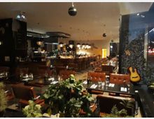 Ein modernes Restaurant mit gedimmter Beleuchtung, lederbezogenen Stühlen und gedeckten Tischen. Im Hintergrund befinden sich eine Bar mit Hängelampen und eine Wand mit schwarzem Mosaik, an der eine Akustikgitarre lehnt.