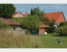 Ein ländlicher Garten mit einer Wiese, überwucherten Sträuchern und Bäumen sowie einem ziegelgedeckten Schuppen aus Holz und Backstein. Im Hintergrund sind Gebäude mit roten Ziegeldächern und eine Metallskulptur erkennbar.